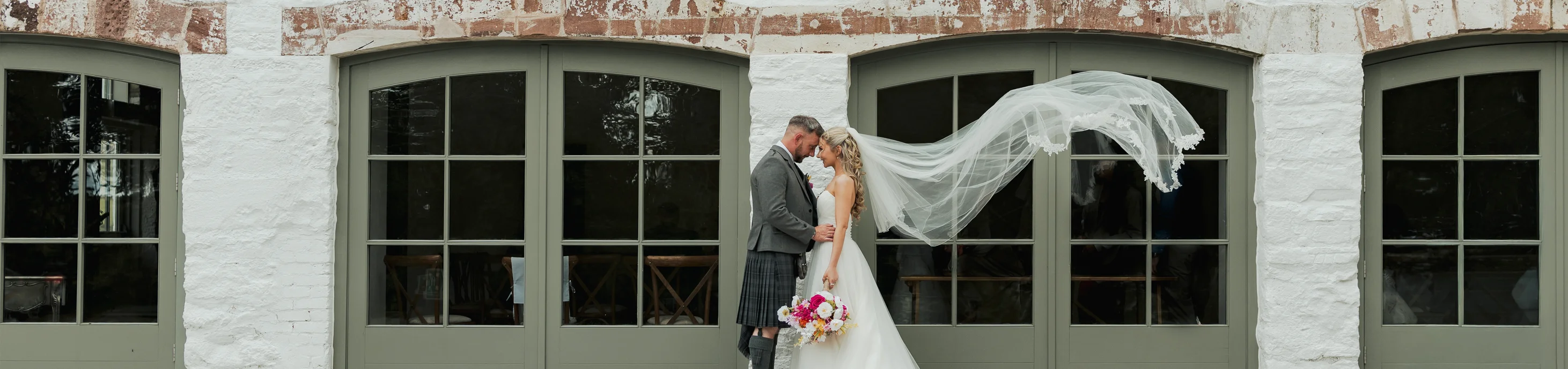 A bride and groom standing together in front of The Arches at Kinclune Steading, with the bride’s veil flowing in the air.
