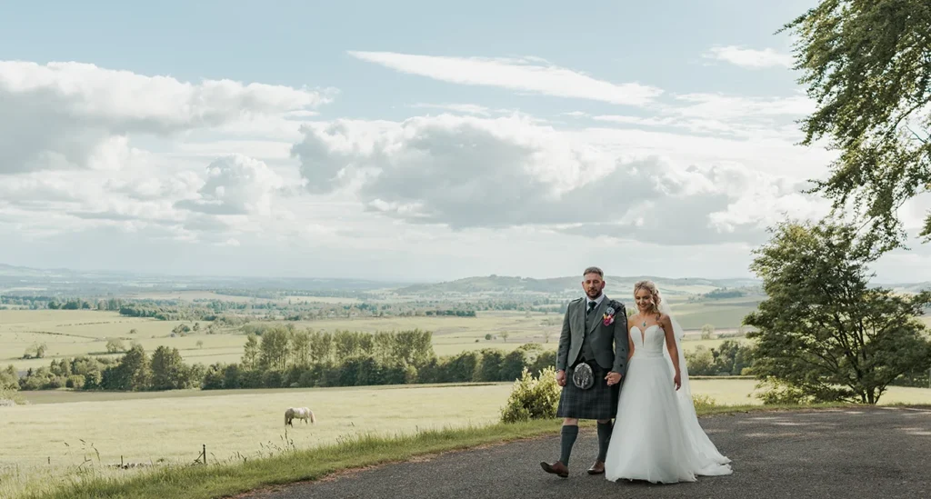 A wedding couple walking hand in hand outside Kinclune House, overlooking the Vale of Strathmore, with a white horse grazing in the field behind them.